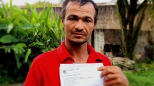 Homem pardo, magro, cabelos curtos, usando camisa vermelha, olha para a câmera exibindo documento