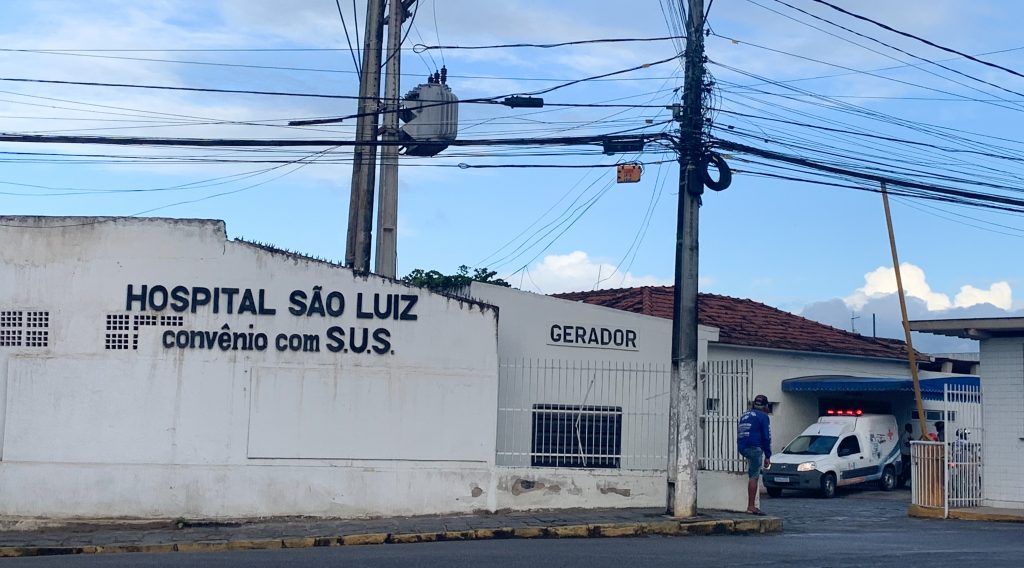 Fachada do hospital São Luiz, com uma ambulância do modelo Fiorino, da Ftat, parada sob um toldo azul em um pátio.