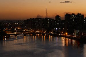 Vista aérea do centro do Recife ao anoitecer, com rio Capibaribe em primeiro plano. Luzes da cidade estão acesas refletindo na água do rio, os contornos dos prédios estão escuros e o céu está alaranjado.