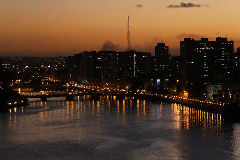 Vista aérea do centro do Recife ao anoitecer, com rio Capibaribe em primeiro plano. Luzes da cidade estão acesas refletindo na água do rio, os contornos dos prédios estão escuros e o céu está alaranjado.