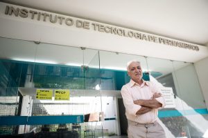 Carlos Wellington: homem branco, idoso, de cabelos brancos ralos, está de braços cruzados com camisa branca e calça bege, em frente à portaria do Instituto Tecnológico de Pernambuco.
