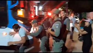 De madrugada, ainda escuro, policiais cercam Aldo Lima (homem pardo, cabelos curtos, jovem vestindo camisa branca e calça jeans), que tenta correr junto a um ônibus com pintura branca e azul.