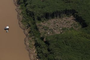 Vista aérea da floresta amazônica, com rio de águas barrentas onde se vê um barco à esquerda da foto e área retangular desmatada à direita da foto.