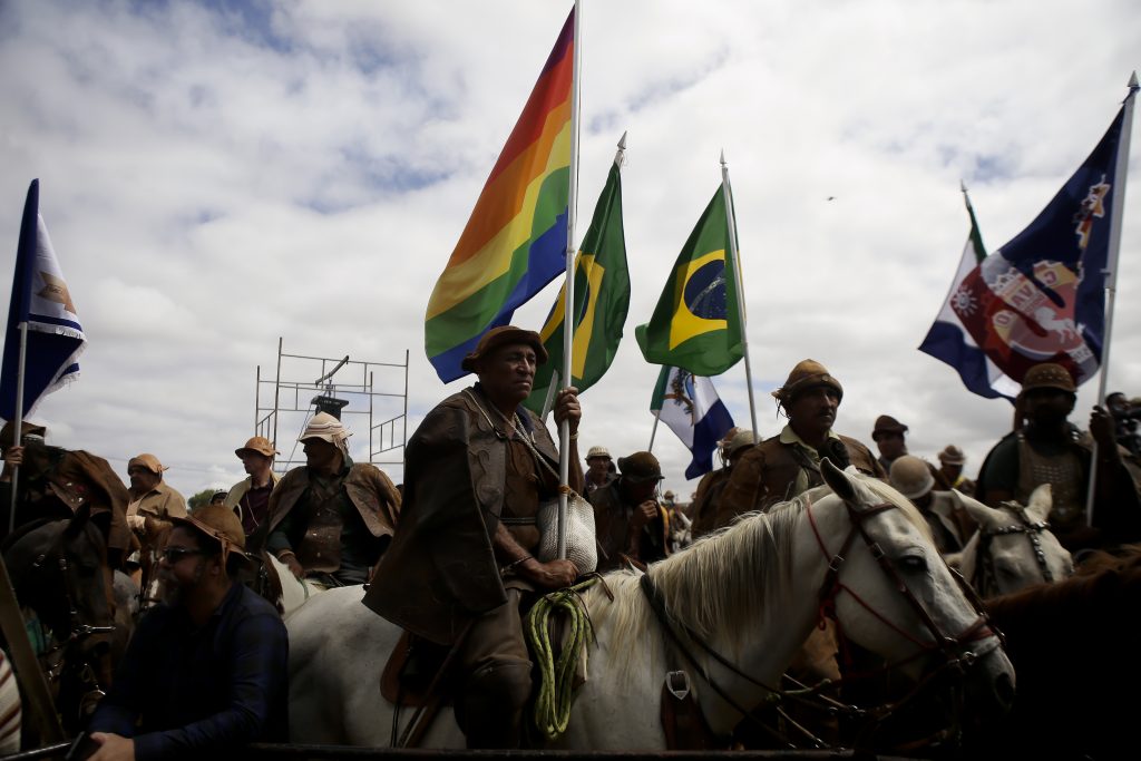 Grupo de vaqueiros montados em seus cavalos e usando trajes de couro, segurando bandeiras do Brasil e uma bandeira com as cores do arco-íris.