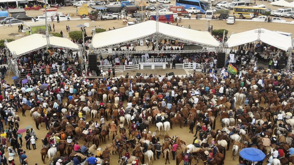 Vista aérea do palco onde se celebra a missa do vaqueiro, com cavaleiros de roupa de couro em seus cavalos, em amplo espaço de terra batida.