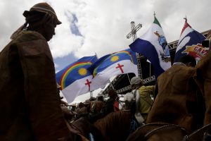 Grupo de vaqueiros em trajes tradicionais de couro, montados em cavalos cujas cabeças aparecem parcialmente na foto, com olhar direcionado para palco que se entrevê entre bandeiras de Pernambuco. Um cruz de estrutura metálica se ergue acima das bandeiras.