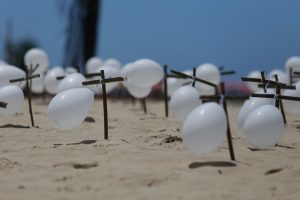 Pequenas cruzes de madeira pintadas de preto, com balões infláveis brancos amarrados, fincadas na areia da praia, em dia de sol sob céu azul.
