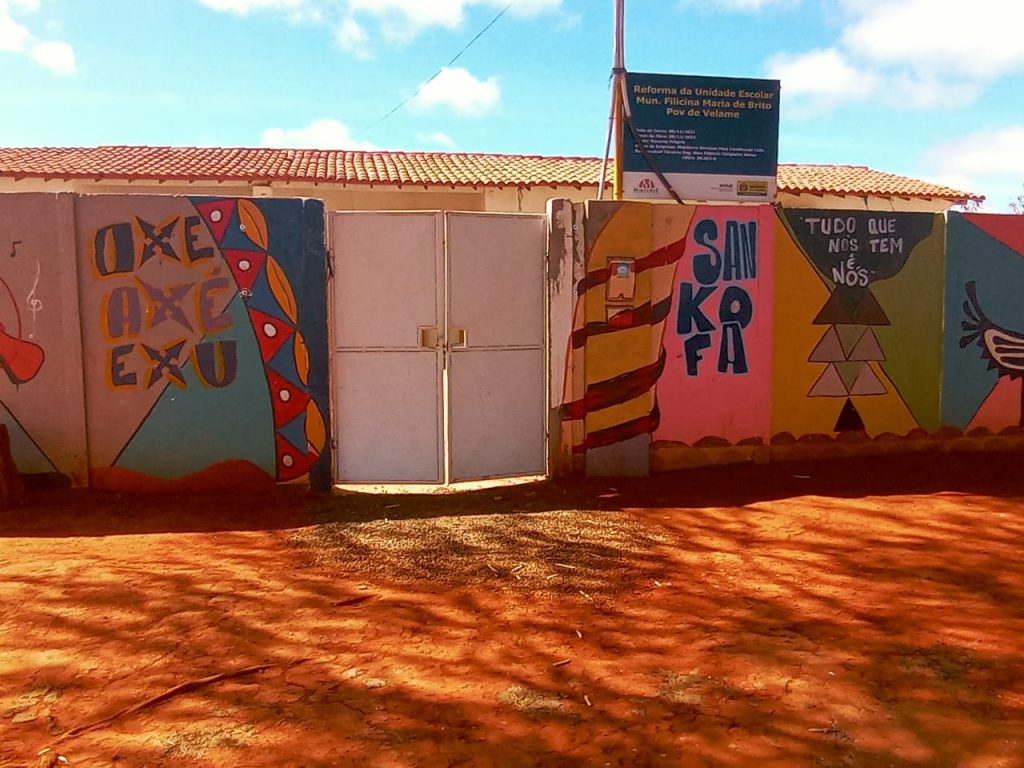 Fachada de escola, com muros pintados, bastante coloridos de azul, amarelo e rosa, onde estão escritas as palavras exu, oxu e axé à esquerda do portão fechado, e, à direita, a palavra sankoka