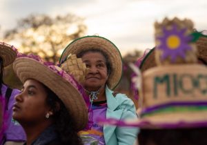 Foto colorida, com foco em uma mulher idosa, usando chapéu de palha e blusa azul clara, com sorriso discreto e olhar para cima, com ar sonhador. Ao redor dela, há silhuetas de outras mulheres com chapéu de palha que estão desfocadas.