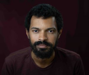 Evandro Cruz Silva: homem negro, jovem, de cabelos e barbas pretas, vestindo camisa marrom e olhando diretamente para a câmera, ao fundo uma parede marrom avermelhada