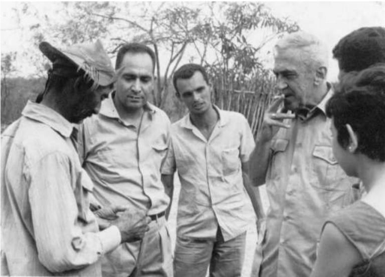 Foto preto e branco no meio da caatinga, em que, numa roda de conversa, um vaqueiro de pele escura e chapéu de couro - à esquerda da foto - conversa com três homens, sendo um deles um idoso de cabelos brancos e uma mulher de cabelos curtos que está de costas, à direita da foto.