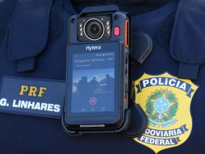 Cãmera de vídeo retangular colocada sobre uniforme preto de policial rodoviária federal cujo rosto não aparece na imagem. à direita da câmera, aparece o emblema da PRF e, à esquerda, o nome PRF Linhares.