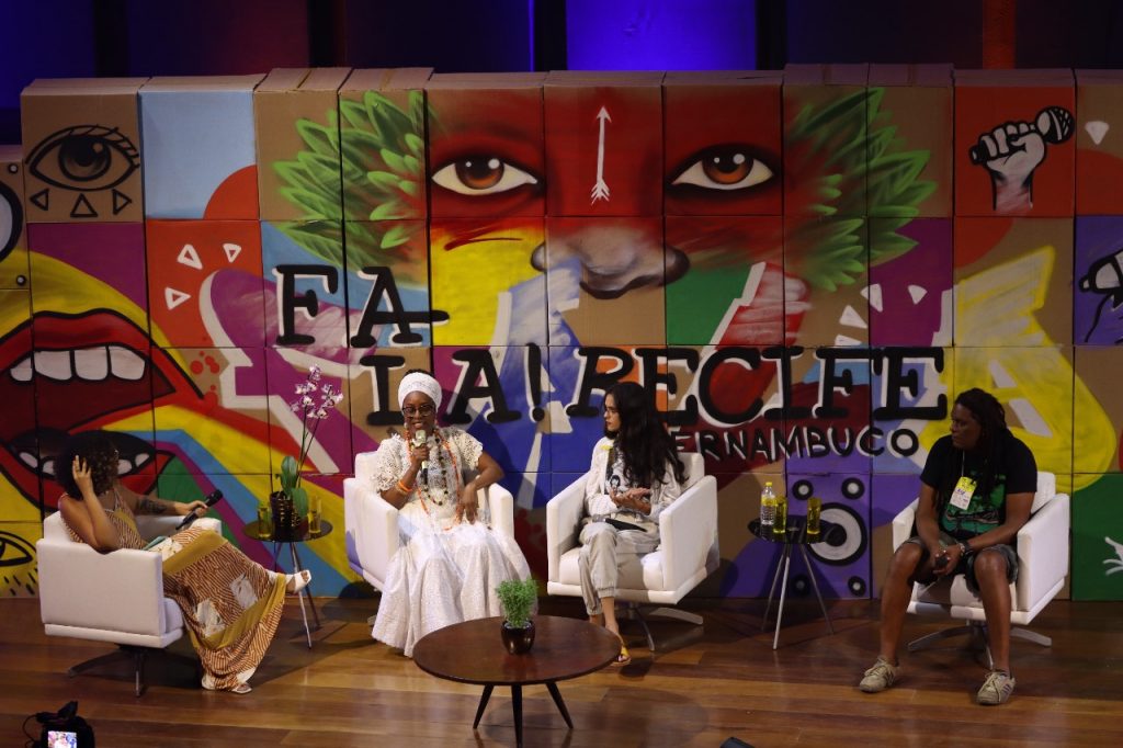 Palco do Festival, com fundo de painel colorido, com quatro pessoas sentadas em poltronas dispostas em semicírculo.