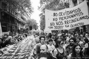 Foto preto e branca de multidão fazendo manifestação em rua de Buenos Aires, carregando um grande mosaico de fotos de vítimas da ditadura e uma faixa com a frase em espanhol Não esquecemos, não perdoamos não nos reconciliamos - são 30.000 - foi genocídio