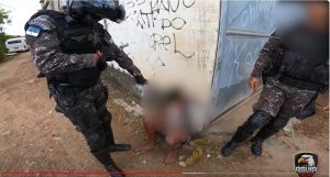 Dois policiais com fardas camufladas em tons de cinza, com capacetes, mantêm rendida uma pessoa cuja imagem está borrada, em um ambiente de aparente pobreza.