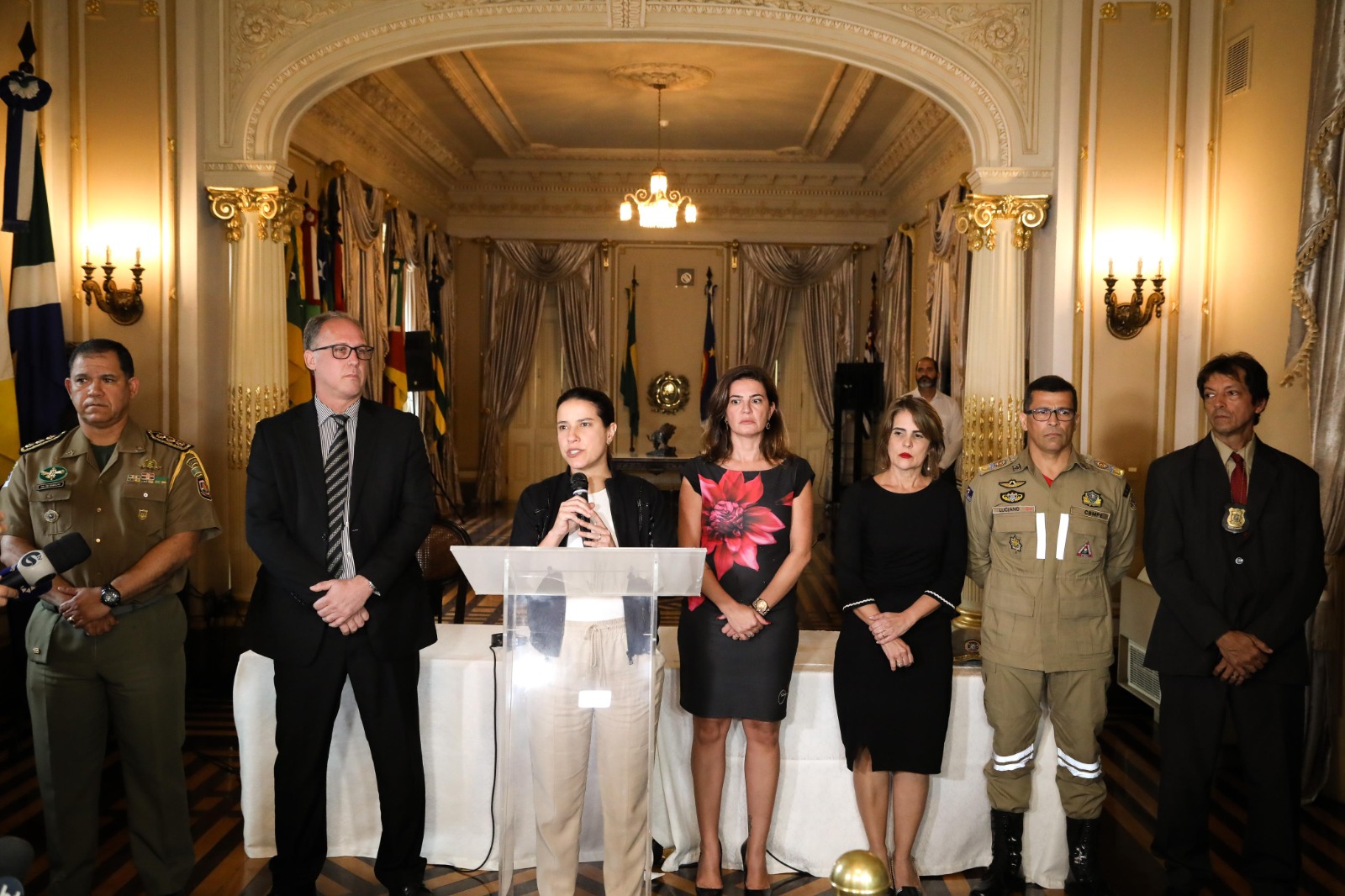 Raquel Lyra, de blusa preta e calça clara, fala ao microfone no em salão nobre do Palácio do Governo, ao lado de autoridades da segurança pública, incluindo duas mulheres de trajes escuros e um policial uniformizado, à sua esquerda, e um homem algo, de meia idade, calvo, com paletó escuro e gravata cinza à sua direita.
