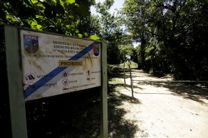 Estrada de acesso à área militar em Suape: em primeiro plano, placa branca, com listas diagonais verde-amarelo desbotada pelo tempo, com a palavra proibido com manchas e riscos. Em segundo plano, estrada de terra, iluminada pelo sol, com vegetação nas duas margens