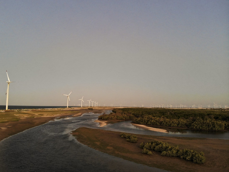 Ao por do sol, imagem de aerogeradores (torres brancas com hélices brancas na parte de cima), produzindo energia eólica em paisagem em faixa de areia no litoral, ao lado do estuário de rio.