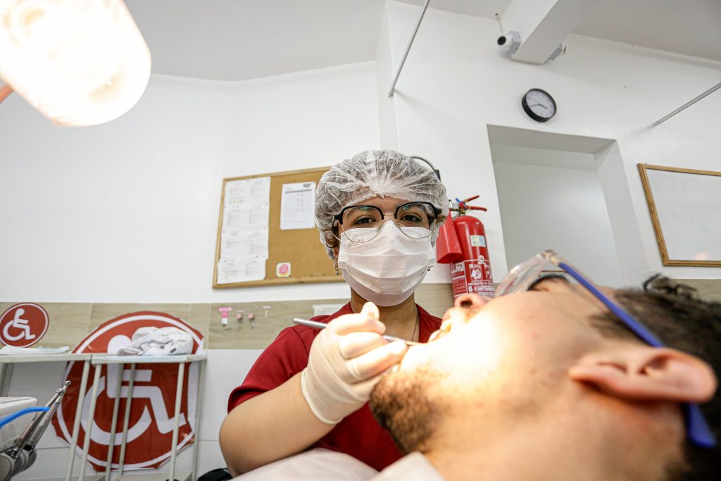 Rebecca Canuto, jovem com máscara cirúrgica e toca, de pele morena, atende paciente odontológico.