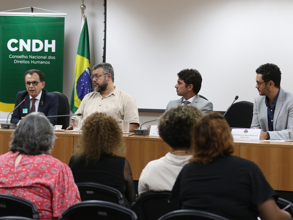 Foto de mesa de trabalhos em auditório, com quatro pessoas sentadas, tendo ao fundo um banner verde com letras brancos sinalizando CNDH - Conselho Nacional de Direitos Humanos.