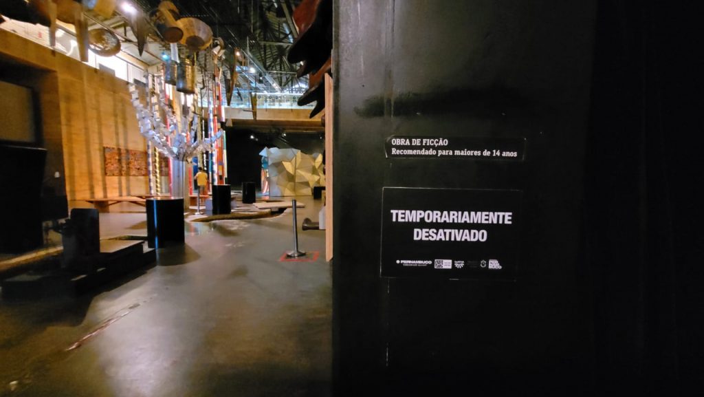 Foto do interior do museu Cais do Sertão, mostrando em primeiro plano uma parede pintada de preto, com um aviso de Temporariamente desativado em letras brancas, tendo sem segundo plano, do lado esquerdo da imagem, peças da exposição.