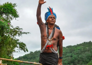 Marcos Xukuru, indígena sem camisa, usando cocar de penas azuis com uma pena vermelha no centro da testa, com colares tradicionais, fotografado ao ar livre, tendo ao fundo uma serra coberta d vegetação