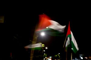 Foto colorida noturna e desfocada de três bandeiras palestinas (pretas, brancas e verdes, com um triângulo vermelho do lado direito, junto ao mastro) tremulando sob céu escuro.