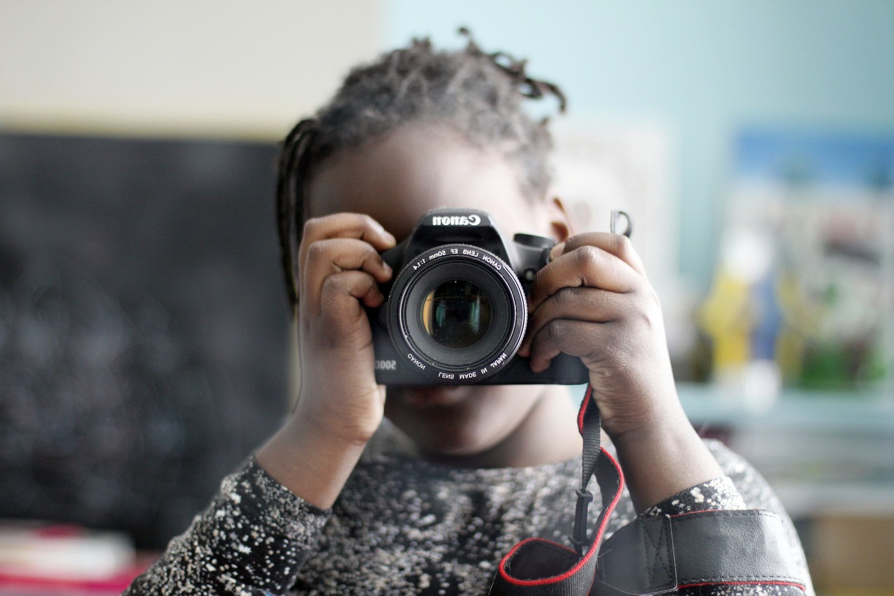 Foto em close de menina negra com o rosto encoberto por uma máquina fotográfica Canon, que ela segura em pose de fotógrafa.