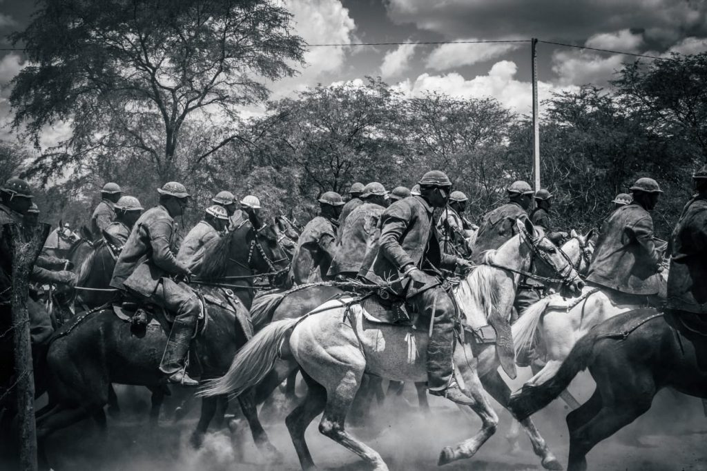 Foto em preto e branco de um grupo de vaqueiros a cavalo passando a galope pela vegetação seca da caatinga.