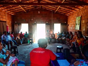 Grupo de quilombolas reunidos em círculo em uma casa de alvernaria, com tijolos sem reboco, e telhado com telhas à mostra. Ao centro, de frente para a janela e de costas para a câmera, no contraluz, está uma mulher de camisa vermelha.