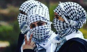 três mulheres jovens, fotografadas do pescoço pra cima, com os rostos cobertos pelo lenço tradicional palestino, branco com estampas em forma de rede na cor preta.