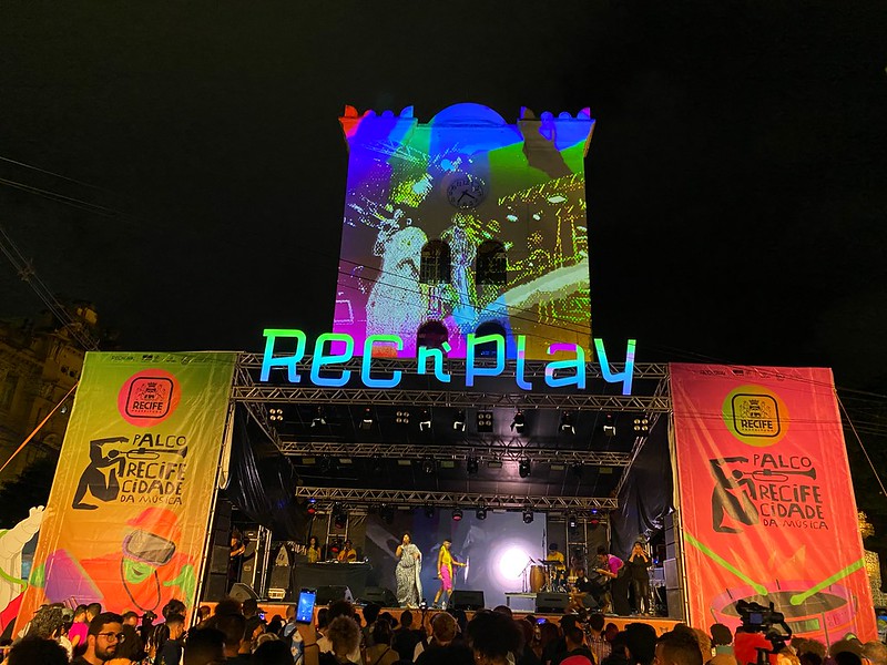 Foto colorida de palco colorido e iluminado, com ao nome rec'n'play no alto do palco, tendo ao fundo os contornos iluminados da Torre de Malakoff