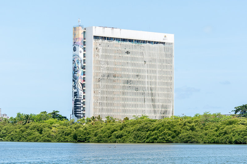 Palácio Capibaribe, edifício-sede da prefeitura do Recife, fotografado à distância em dia de céu azul sem nuvens, tendo o rio Capibaribe e o manguezal na parte inferior da imagem