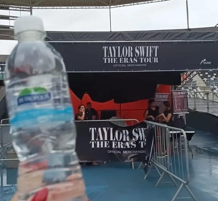 em primeiro plano, mão de mulher segura garrada plástica de água mineral em frente à entrada do show de Talyor Swift The Eras, na frente do estádio Engenhão