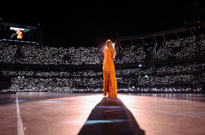 foto colorida de Taylor Swift, em pé sobre o palco, usando um vestido longo de cor laranja, de costas para a câmera e em frente a uma multidão que lota as arquibancadas de um estádio à noite, com luzes dos celulares se destacando nas arquibancadas.