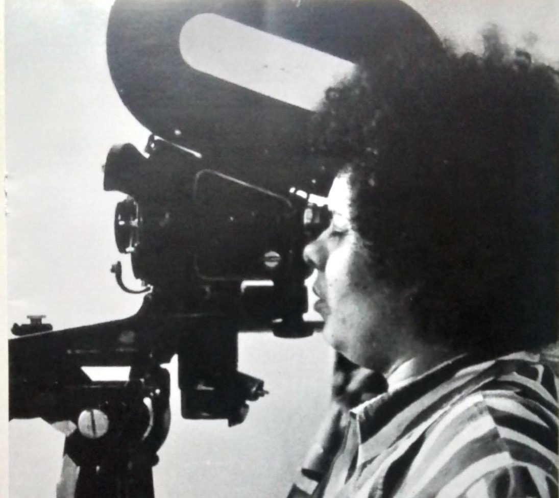 Foto em preto e branco de Adélia Sampaio: mulher negra, de cabelos cortados no estilo black power, usando camisas de listas verticais, olhando para visor de antiga câmera de cinema.