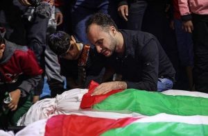 Homem chora, debruçado sobre cadáver de parente envolto em tecido branco, verde e vermelho, cores da bandeira palestina. Ele é observado de perto por um menino de cabelos pretos e pele morena.