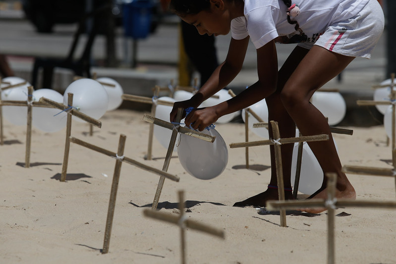 Criança negra, de short e camiseta branca, amarrando balões de ar brancos em cruzes de madeiras fincadas na areia da praia.