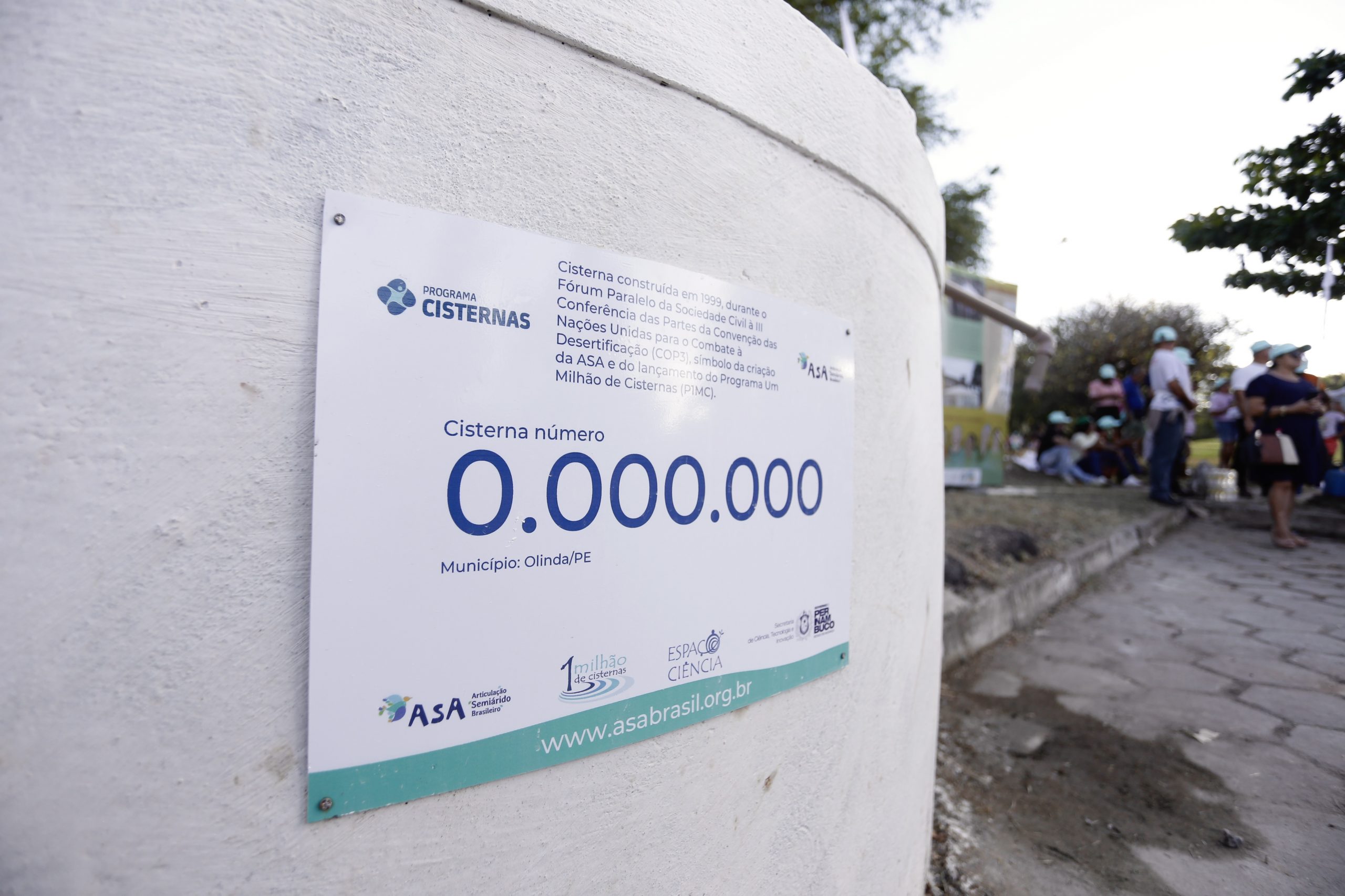 Foto colorida de detalhe de cisterna de cor branca, com placa do Programa Um Milhão de Cisternas indicando o número 0.000.000