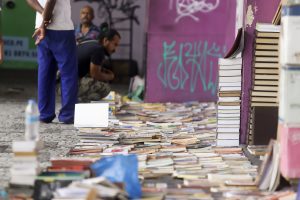Em primeiro plano, livros espalhados por uma calçada, com um homem de barba e camisa preta agachado olhando o que está à venda.