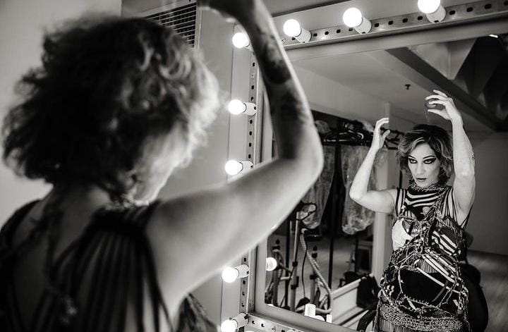 Foto em preto e branco de Karina Buhr, maquiada pronta para entrar em cena, se olhando em um espelho no camarim.