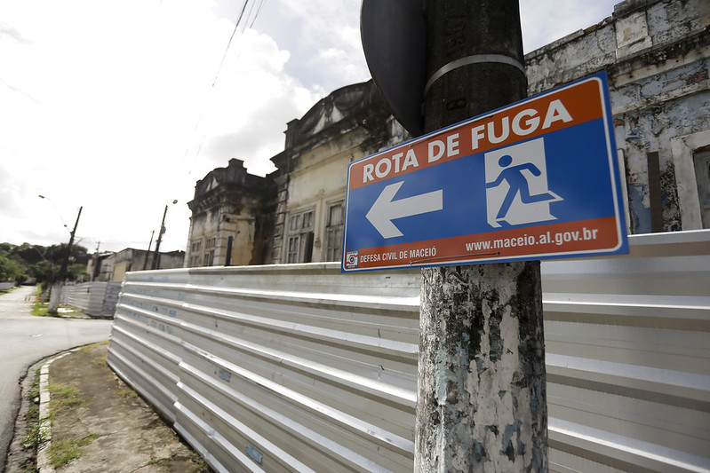 Em primeiro plano, à direita da imagem, pequena placa azul e vermelha com seta branca sinalizando rota de fuga, em frente a uma fachada de imóvel com tapumes de alumínio.