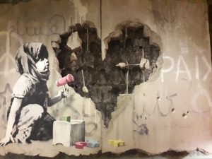 foto colorida de intervenção artística no muro cinzento que separa Israel da Cisjordânia. A foto mostra o desenho de uma menina em preto e branco ao lado de um coração escavado no muro.