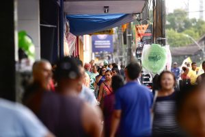 Multidão transitando por uma calçada em rua de comércio varejista em Recife, com o rosto das pessoas que estão em primeiro plano desfocados.