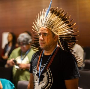 Foto colorida de Dinanam Tuxá durante a COP28, usando cocar tradicional do povo indígena tuxá e colares nas cores azul e vermelha. Ele está de camisa preta em pé num auditório de pares marrons, em meio a outras pessoas sentadas, fora de foco.