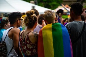 Três pessoas vestidas de maneira informal observam uma multidão de costas para a câmera. Uma delas, de cabelo curto á direita da imagem, está usando uma bandeira do arco-íris nas pendurada sobre os ombros.