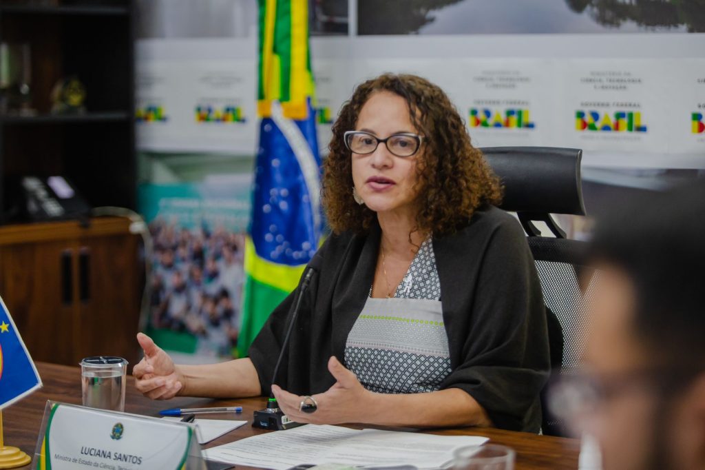Foto colorida de Luciana Santos (mulher negra, de cabelos cacheados e óculos de aro escuro), por trás de uma mesa e à frente de uma bandeira do Brasil. Ela usa blusa de listas cinzas escuro e cinza claro, com um casaco preto por cima dos ombros.