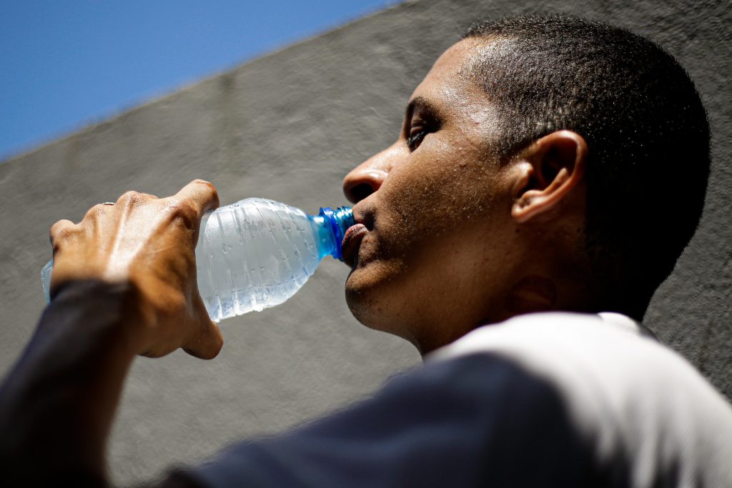 Foto colorida de homem jovem negro, de perfil, bebendo água direto de uma garrafa de plástico azul transparente.