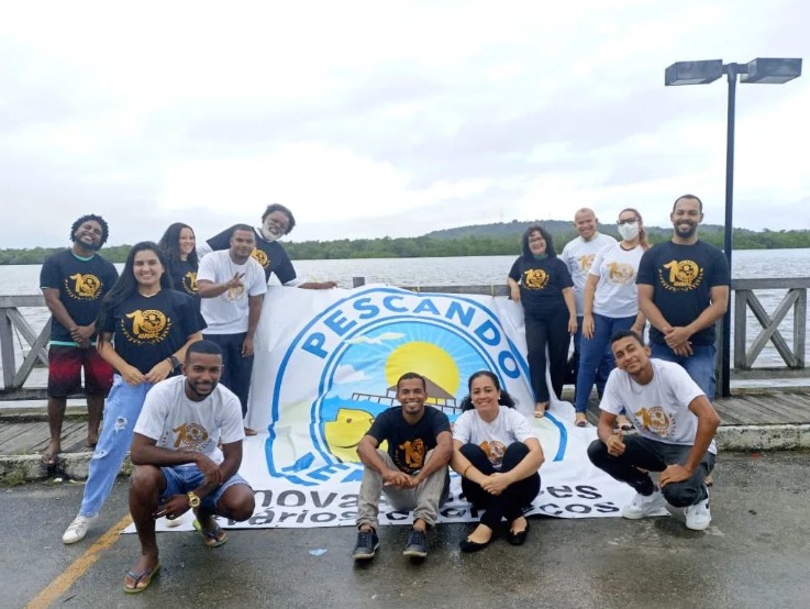 Foto colorida de grupo de 13 pessoas ao redor de uma grande banner com a marca do projeto Pescando Memórias com orla de rio ao fundo