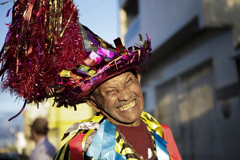 Foto colorida de Mestre Martelo: ele é um homem negro, idoso, de sorriso escancarado, usado um chapéu enfeitado de miçangas e fitas, usando uma fantasia típica feita de retalhos de tecido colorido.